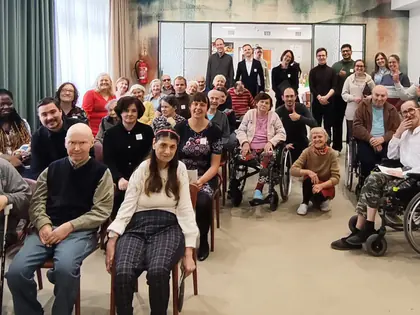 Grupowe zdjęcie mieszkańców, gości i pracowników na sali, część siedzi na krzesłach i wózkach
