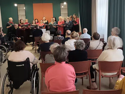 Chór w czerwonych i czarnych strojach śpiewa przed publicznością siedzącą na krzesłach