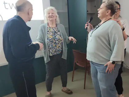 Dwoje starszych ludzi tańczy, obok kobieta z mikrofonem, w tle biały ekran z tekstem piosenki