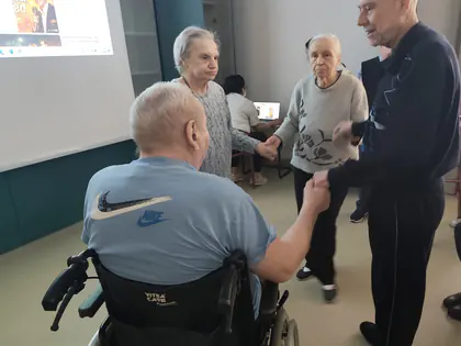 N zdjęciu mężczyzna na wózku inwalidzkim trzyma starsze osoby za rękę w kręgu tanecznym