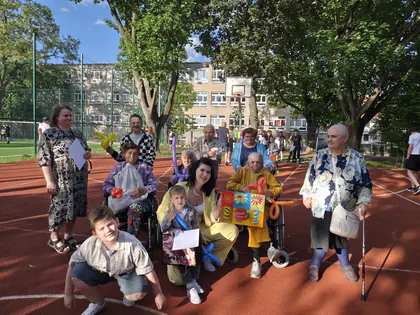 Grupa młodzieży z seniorami pozuje do zdjęcia, z przodu kobieta z dzieckiem trzyma kolorowy obraz