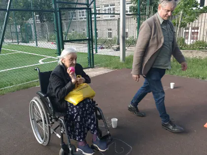 Po lewej starsza kobieta na wózku trzyma piłeczkę do gry, z prawej idzie w jej stronę mężczyzna