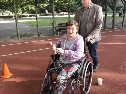 Mężczyzna pcha wózek, na którym siedzi kobieta, w tle boisko szkolne