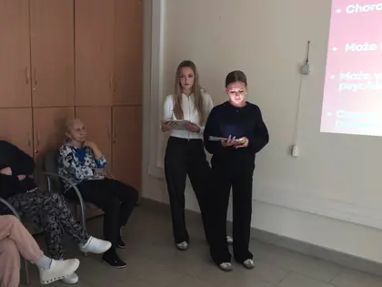 Dwie studentki prowadzą wykład, czytając z tabletów. Obok na krzesłach siedzą seniorzy, na ścianie wyświetlany jest slajd