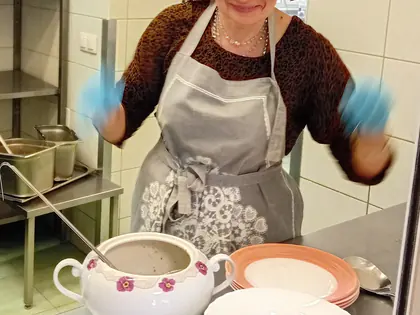 Radosna kobieta w średnim wieku w chustce i fartuchu, przed nią waza z zupą i zastawa stołowa, w tle kuchnia