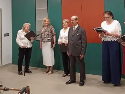 Grupa śpiewaków daje koncert, starsza pani po lewej i młodsza po prawej trzymają czarne teczki z tekstami piosenek, w tle kolorowe meble