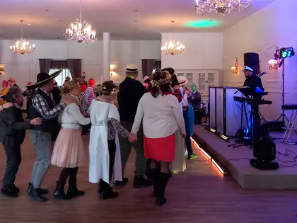 Uczestnicy balu w przebraniach tańczą pociągiem na sali z żyrandolami, w tle DJ przy stanowisku muzycznym