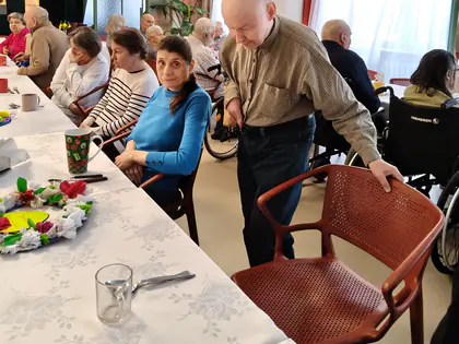 Pan w średnim wieku siada przy długim stole z białym obrusem i zastawą, obok siedzą inne osoby