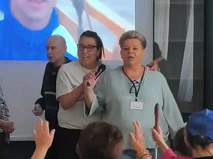 Dwie panie z mikrofonem rozbawiają starsze osoby dookoła, z przodu kobieta w żółtej bluzce tańczy z podniesionymi rękami