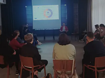 Grupa osób siedzi w kręgu na krzesłach, z przodu sali ekran ze slajdem, kobieta w czerwonej marynarce czyta słuchającym uczestnikom